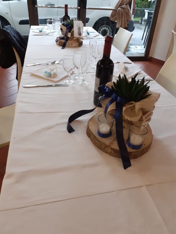 Apparecchiatura della tavola: tovaglia bianca, bottiglie di vino, bicchieri, candele e composizione di piante decorative su una base di legno.