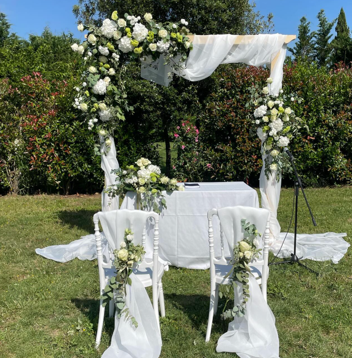 Arco nuziale e sedie decorate con fiori bianchi e tessuto trasparente, immersi in un giardino soleggiato.