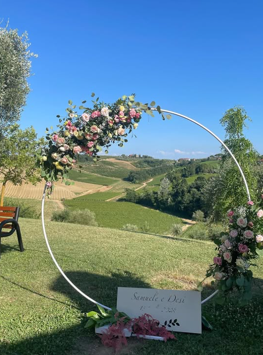 Arco nuziale circolare bianco con fiori, insegna e campagna toscana sullo sfondo.