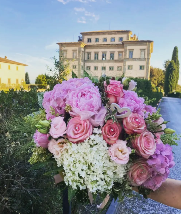 Un mazzo di fiori rosa e bianchi tenuto davanti a un grande edificio con un cielo azzurro.