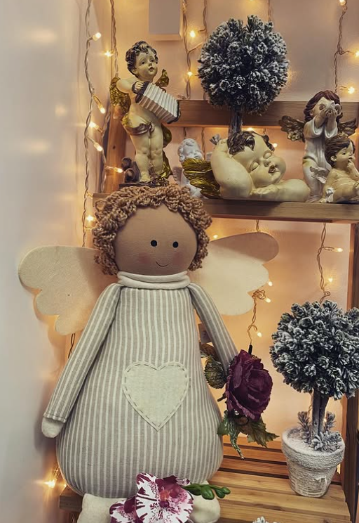 Bambola di stoffa a forma di angelo con le ali, che tiene in mano una rosa, davanti a uno scaffale con statuette di angeli e luci decorative.