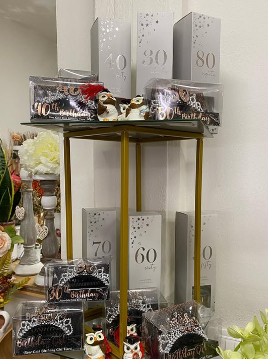 Scaffali che espongono regali di compleanno, tra cui numeri in scatola (50, 60, 70, 80), dolcetti al cioccolato e statuette di gufi.