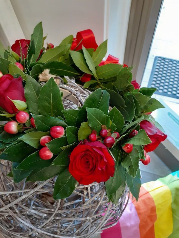 Corona di rose rosse, foglie verdi e bacche su un cesto intrecciato, accanto a una finestra.