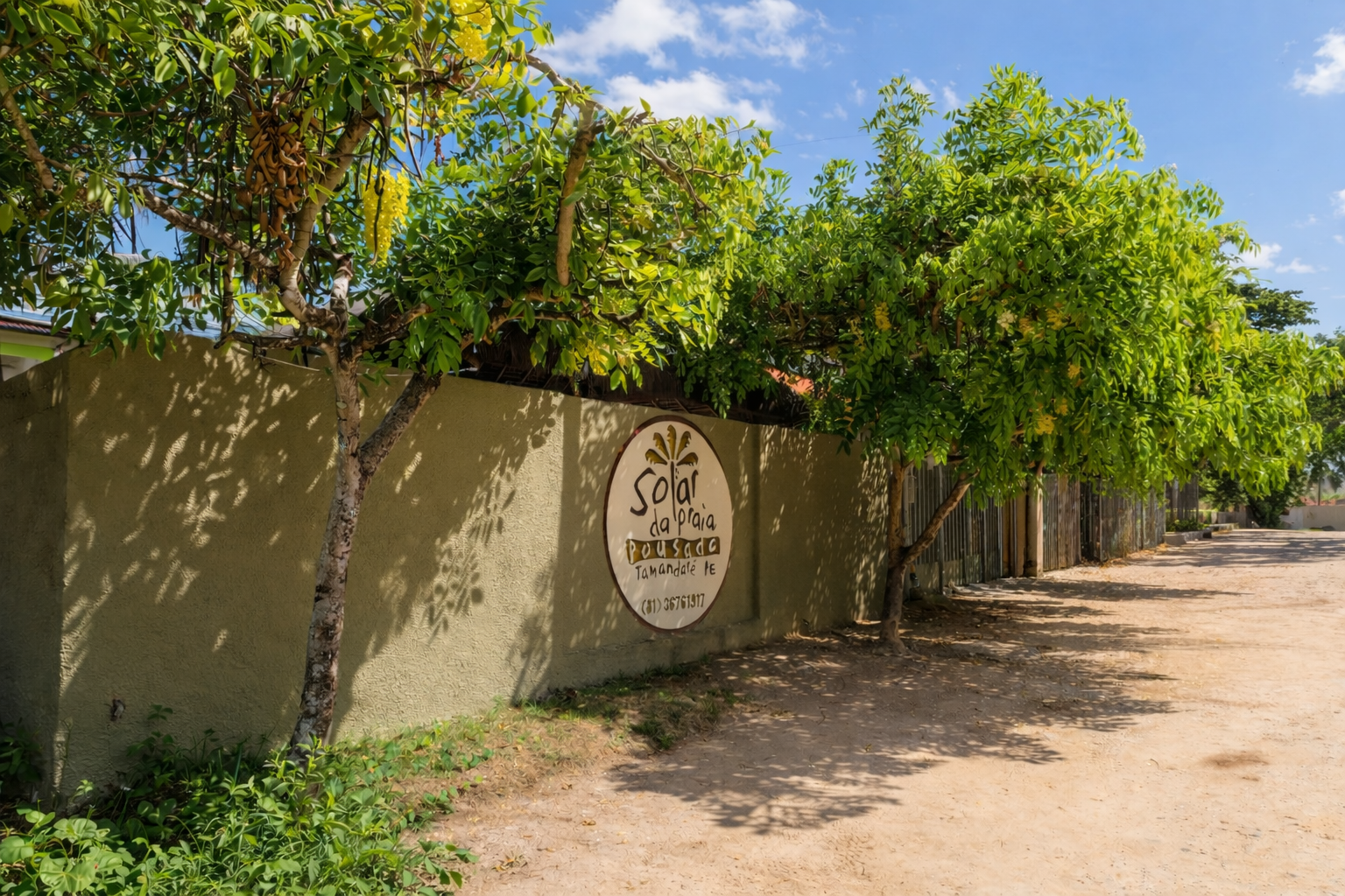 Uma parede de estuque com uma placa parcialmente encoberta por folhagem verde, ao lado de uma estrada arenosa.