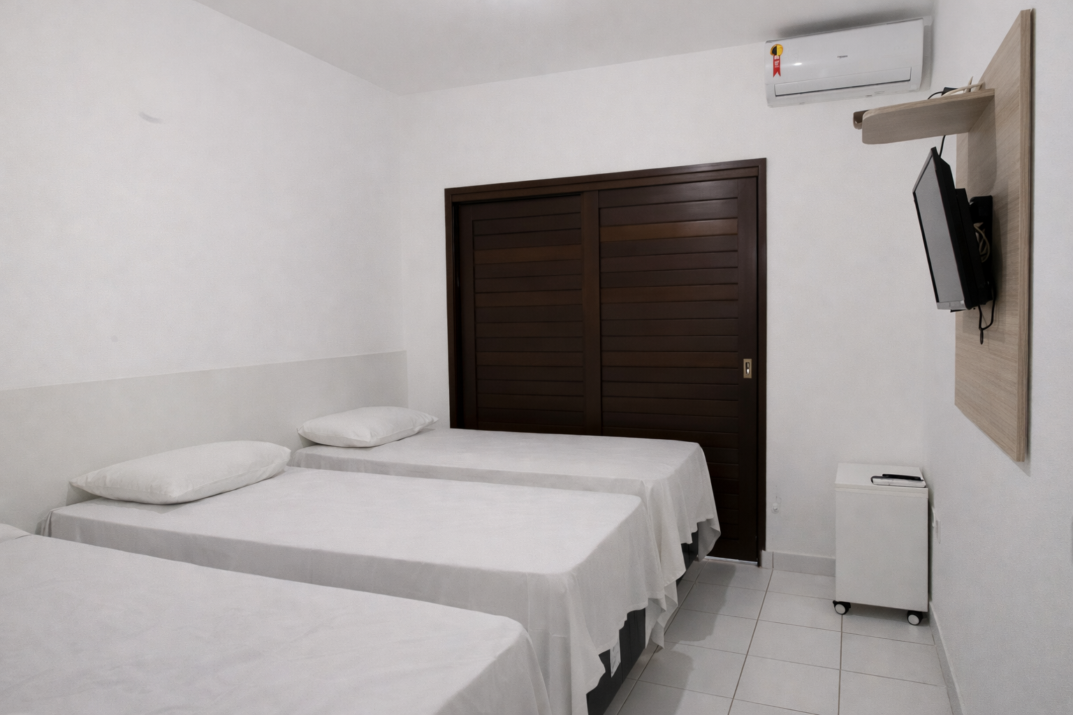 Quarto com três camas de solteiro, armário de madeira escura, TV fixada na parede, ar condicionado e paredes brancas.