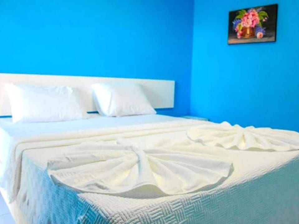 Cama branca com toalhas dobradas, contra paredes azuis com um quadro floral.