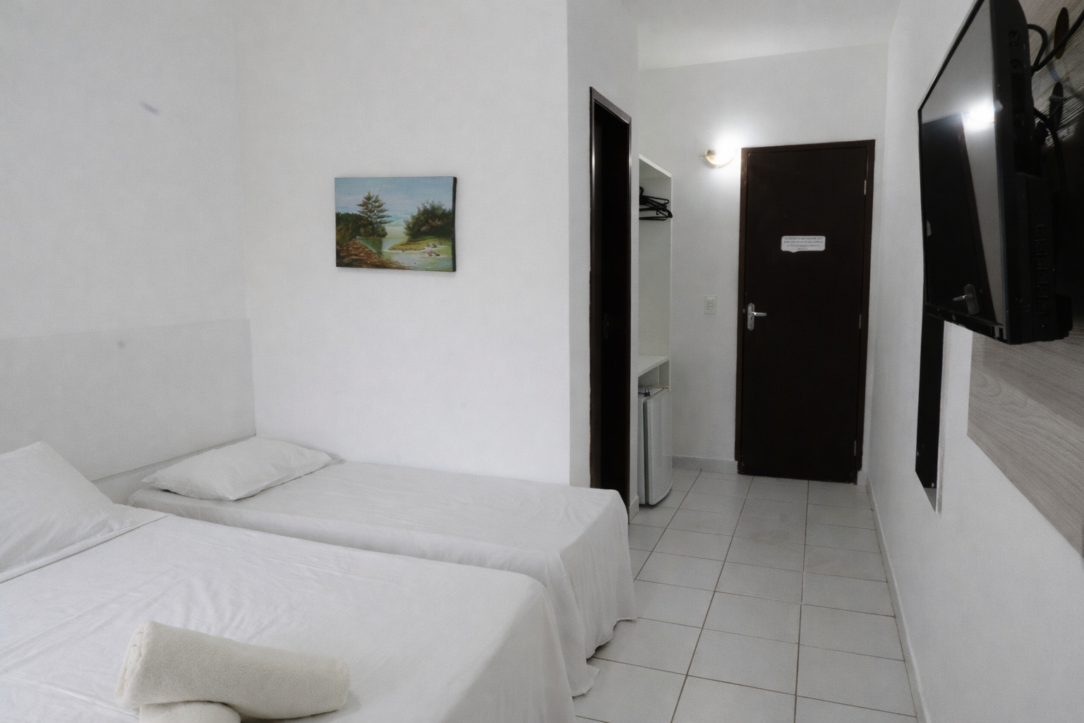 Um pequeno quarto de hotel branco com duas camas de solteiro, uma televisão e uma porta que dá para um corredor.