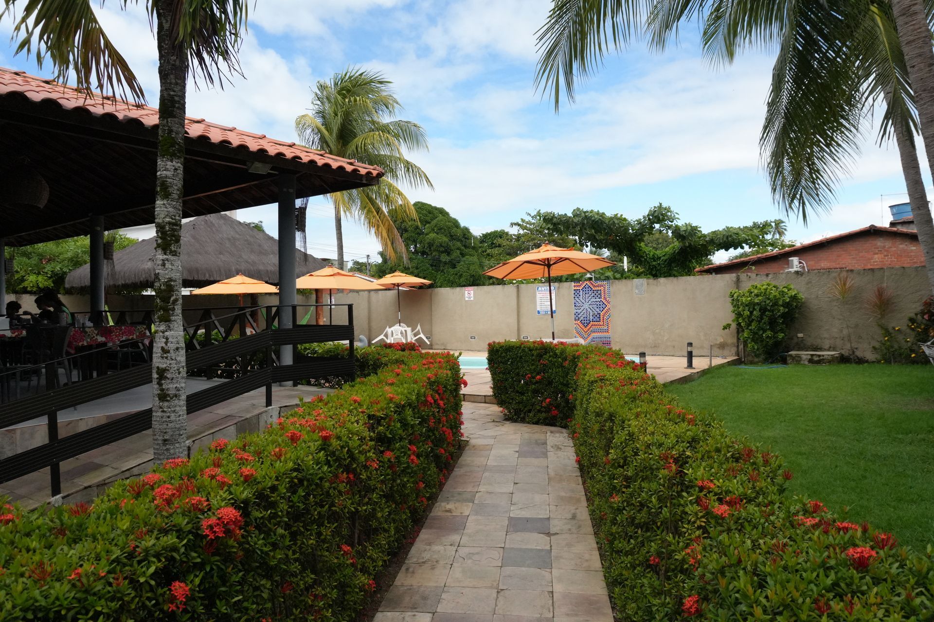 Caminho que leva a uma piscina ladeada por sebes floridas de vermelho, com um pátio coberto à esquerda e palmeiras acima.
