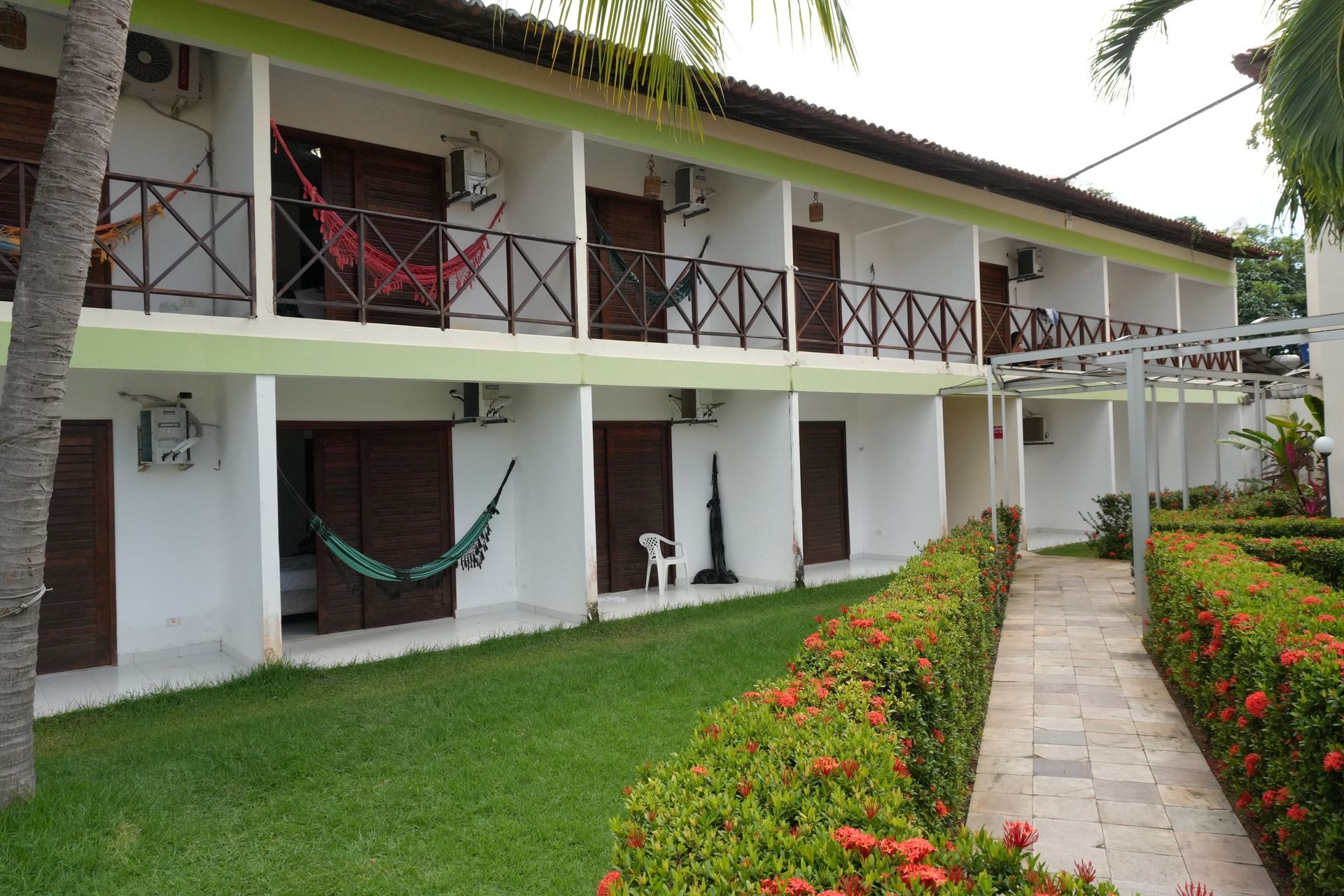 Exterior de um prédio branco de dois andares com portas marrons, varandas e detalhes em verde. Uma passarela e cercas vivas acompanham o edifício.