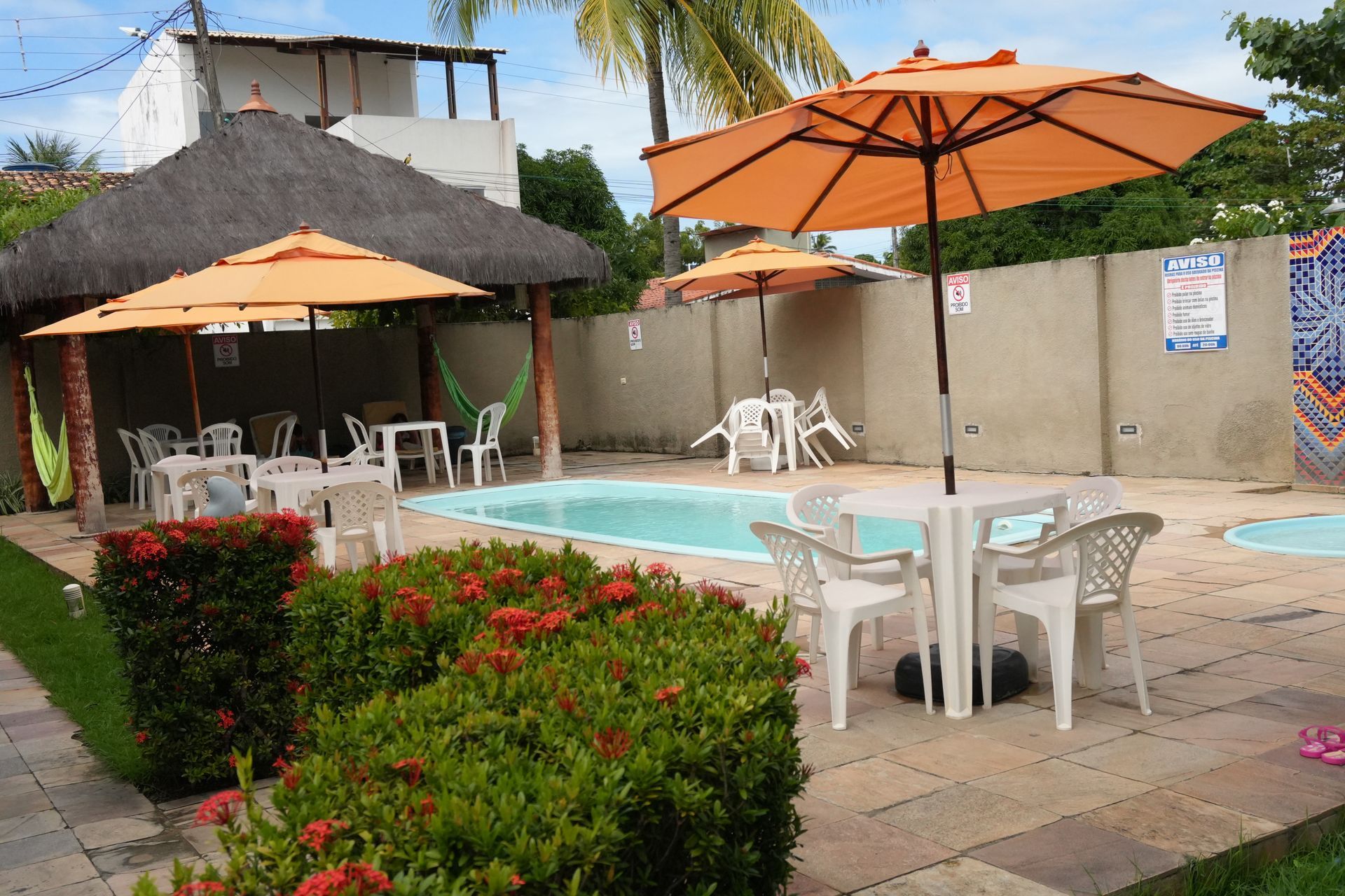 Área da piscina com mesas, guarda-sóis e uma estrutura com teto de palha. Vegetação exuberante e uma pequena piscina também são visíveis.