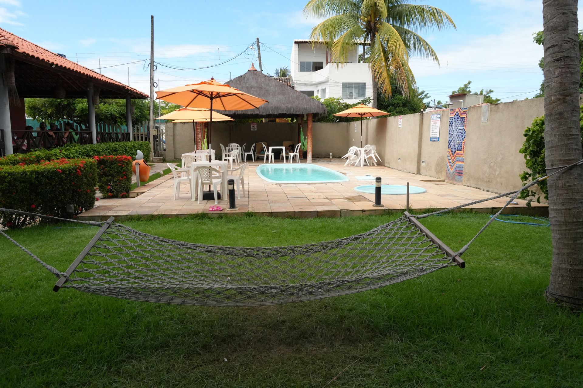 Rede em frente à piscina. Guarda-sóis laranja, bar tiki, mesas e cadeiras brancas num dia ensolarado.