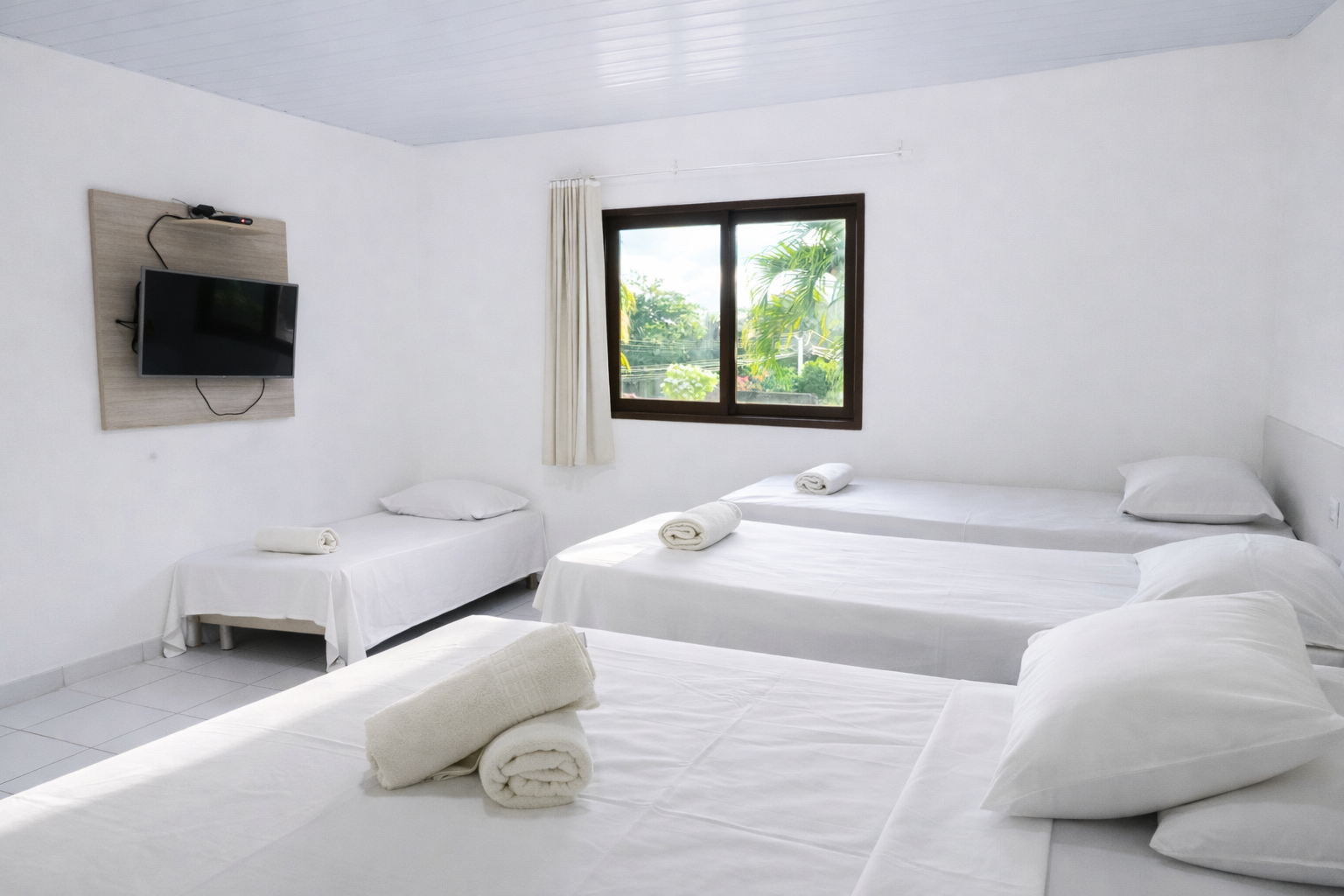 Quarto com três camas e uma pequena TV na parede. O quarto é branco e tem uma janela.