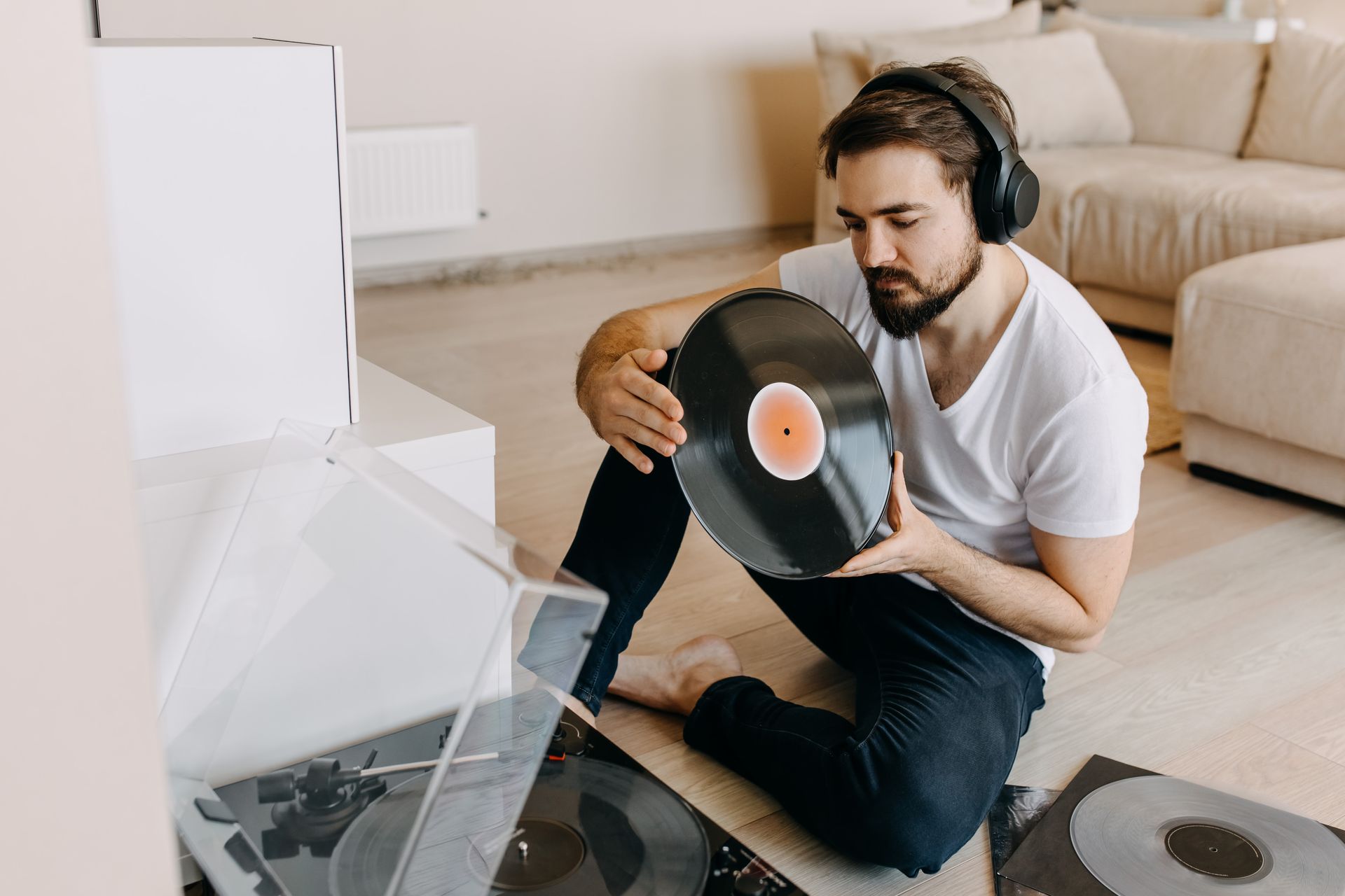 Man holding vinyl record, sitting by record player, wearing headphones. Interior.