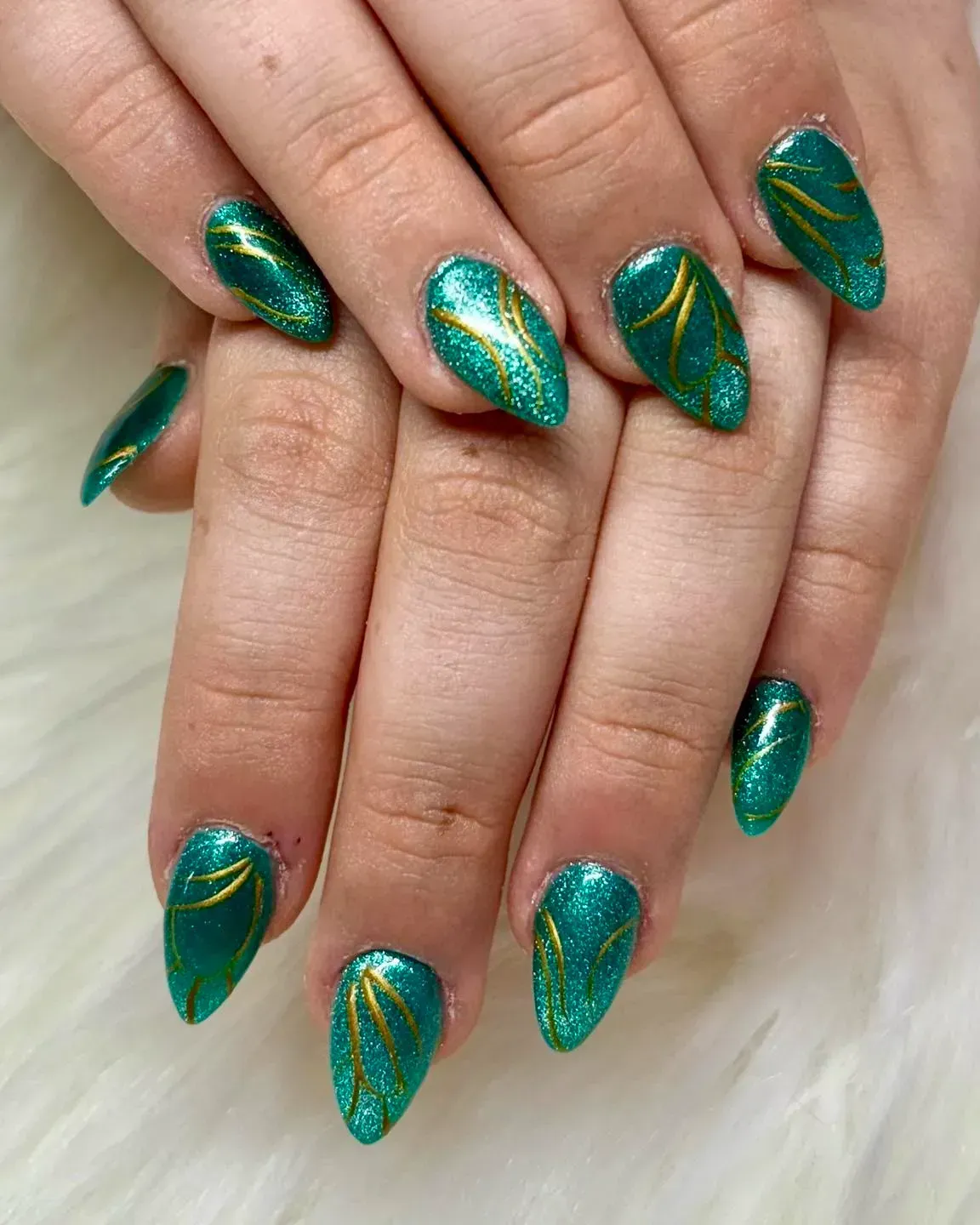 Hands with almond-shaped teal nails. Gold glitter accents in curved lines.