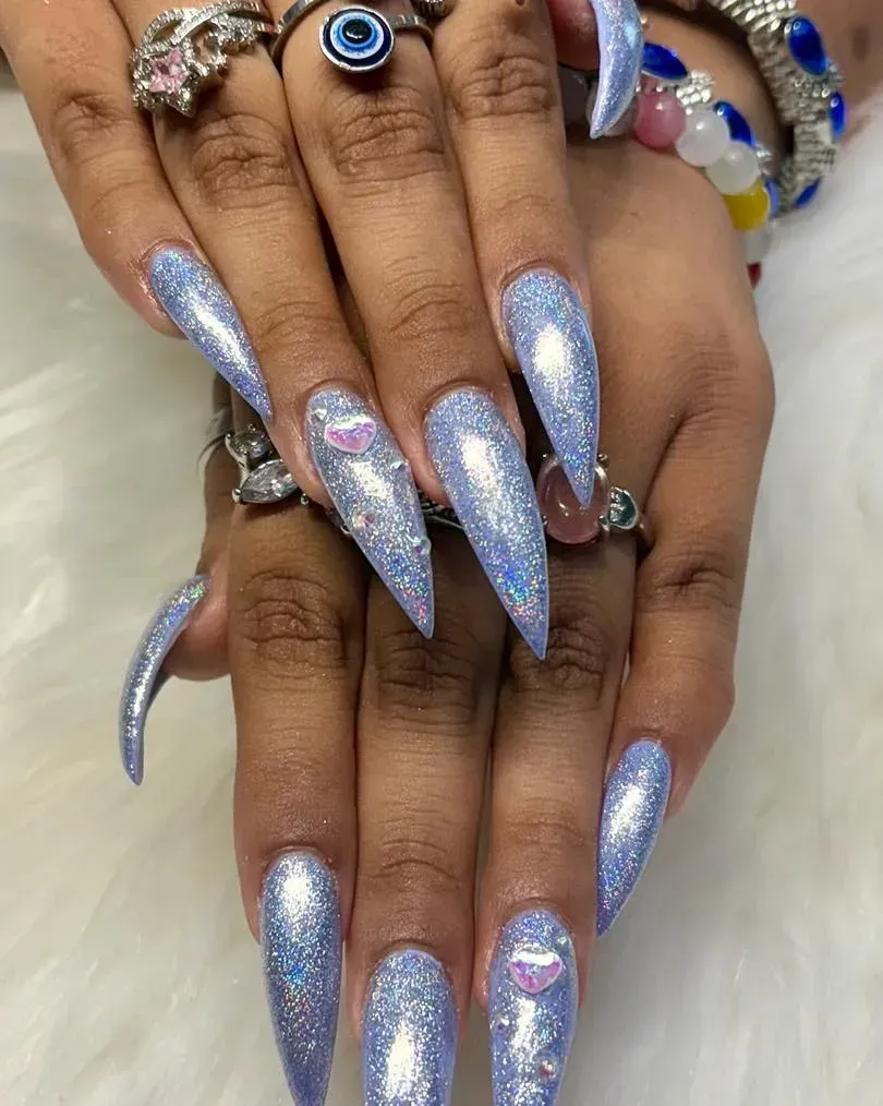 Long, pointed nails with blue glitter, some adorned with small lip-shaped decorations, resting on a white surface.