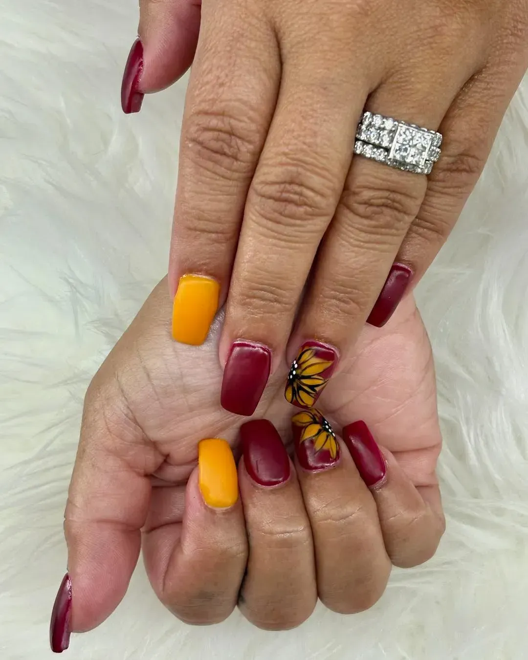 Hands with long square nails: burgundy and yellow. One nail features a sunflower and butterfly.