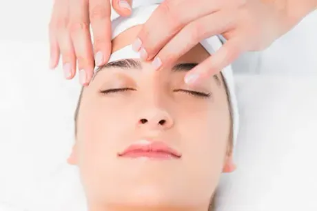 Woman receiving eyebrow waxing treatment. Hands holding a wax strip to her forehead, eyes closed.