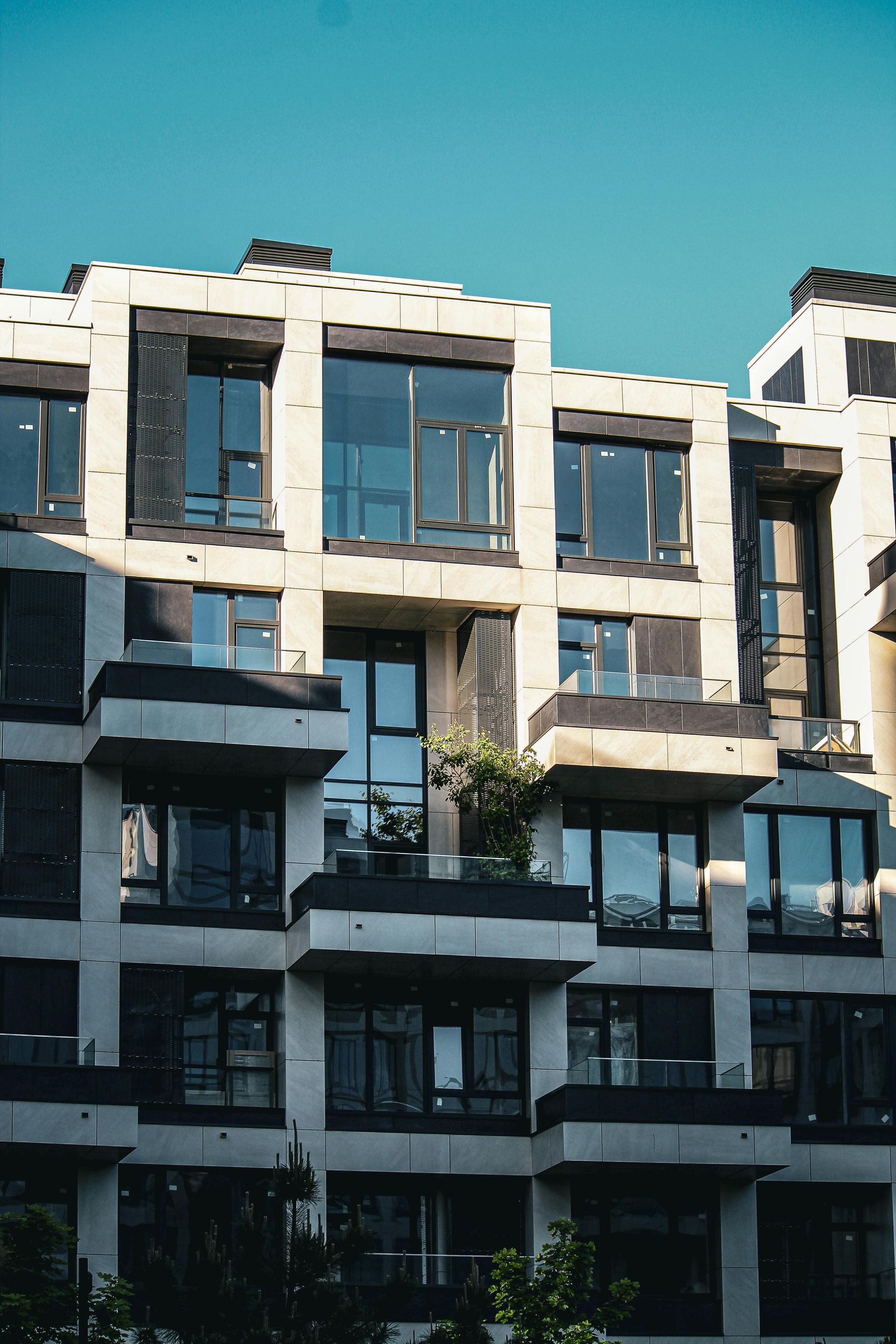 A modern, tan multi-story apartment building with balconies and a glass-fronted ground floor under a clear blue sky.