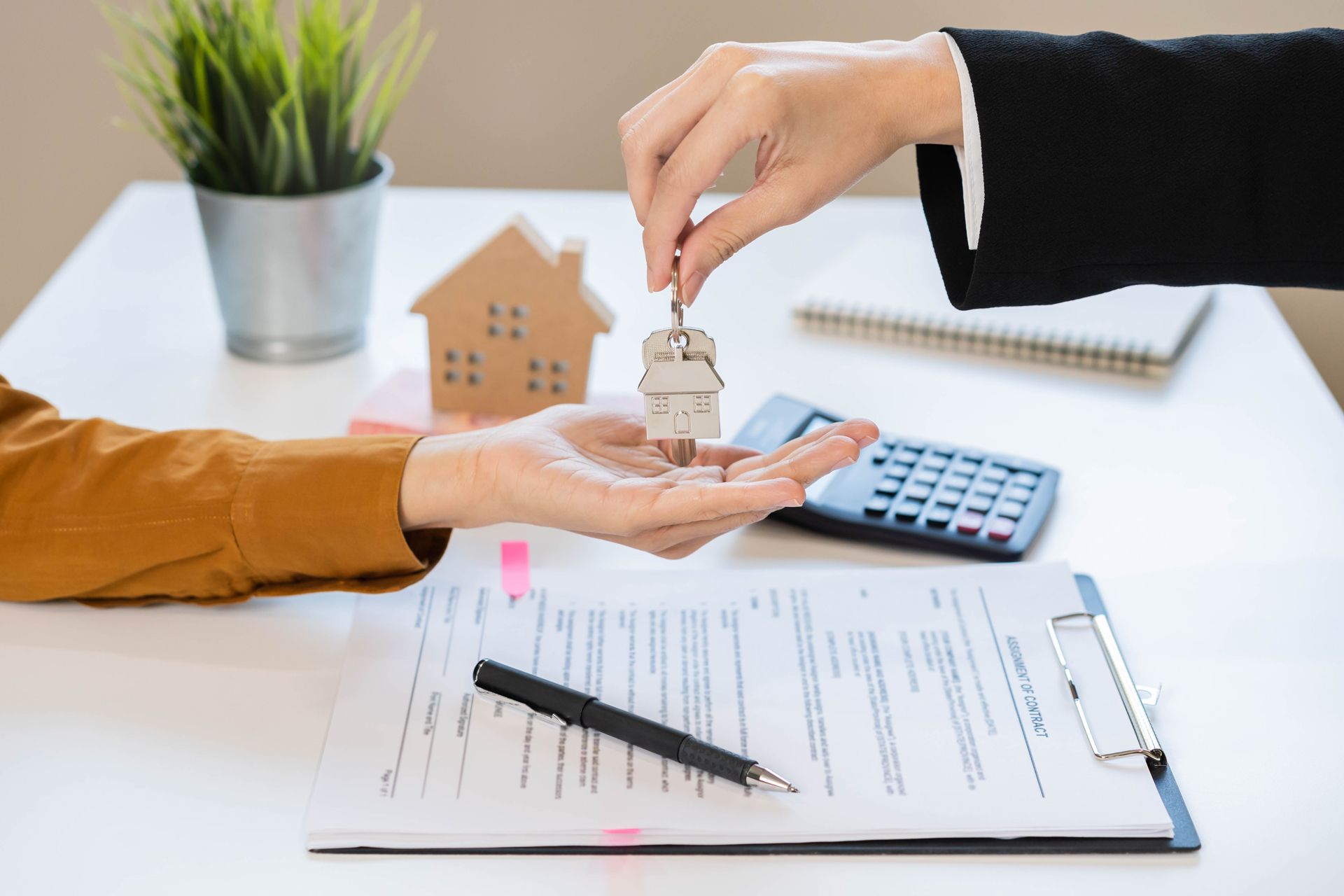 A hand hands house keys to another person over a contract on a desk with a small model house, calculator, and pen.