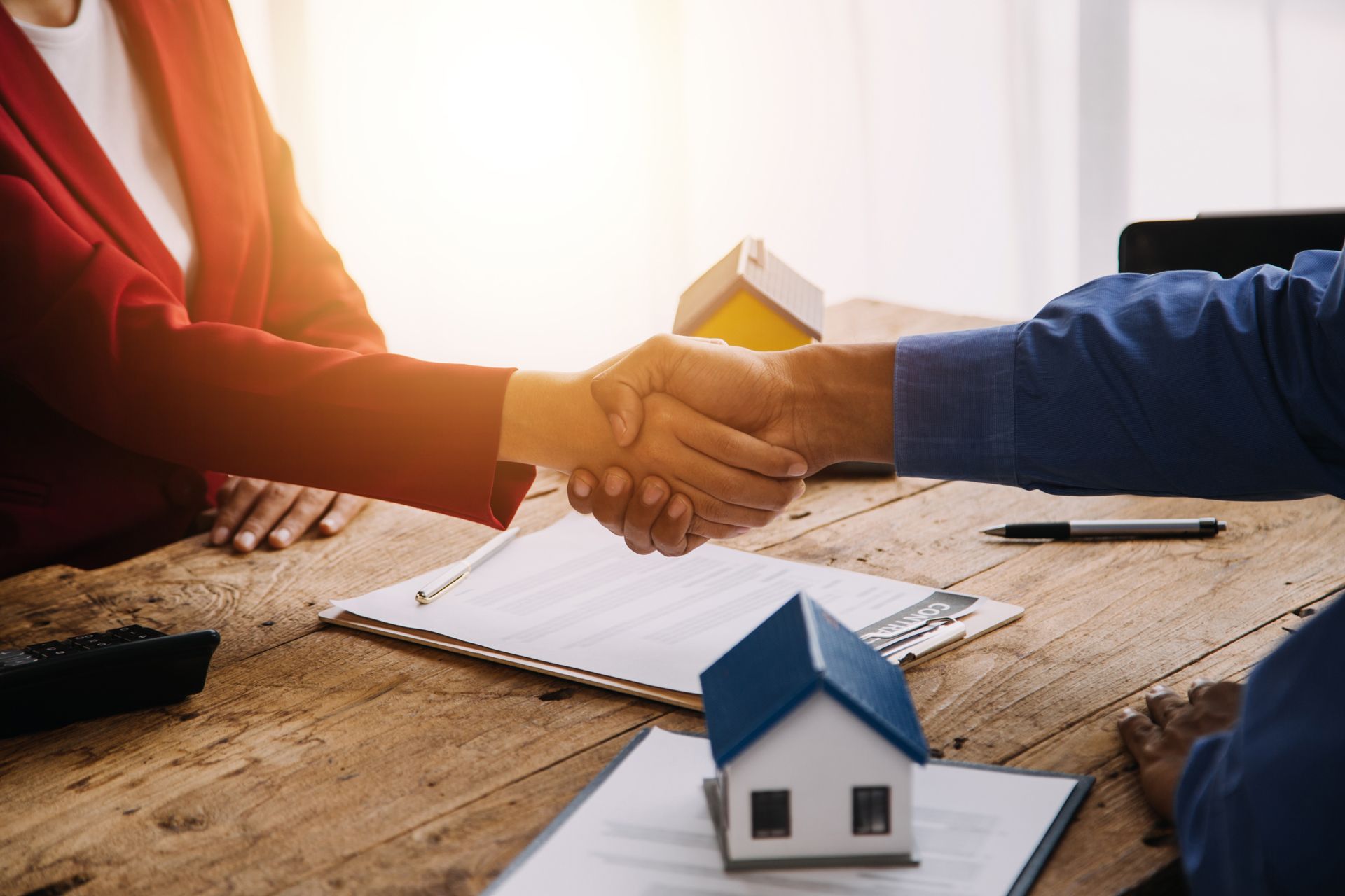 Two people shaking hands over a wooden desk with house models and contracts, representing a real estate agreement.