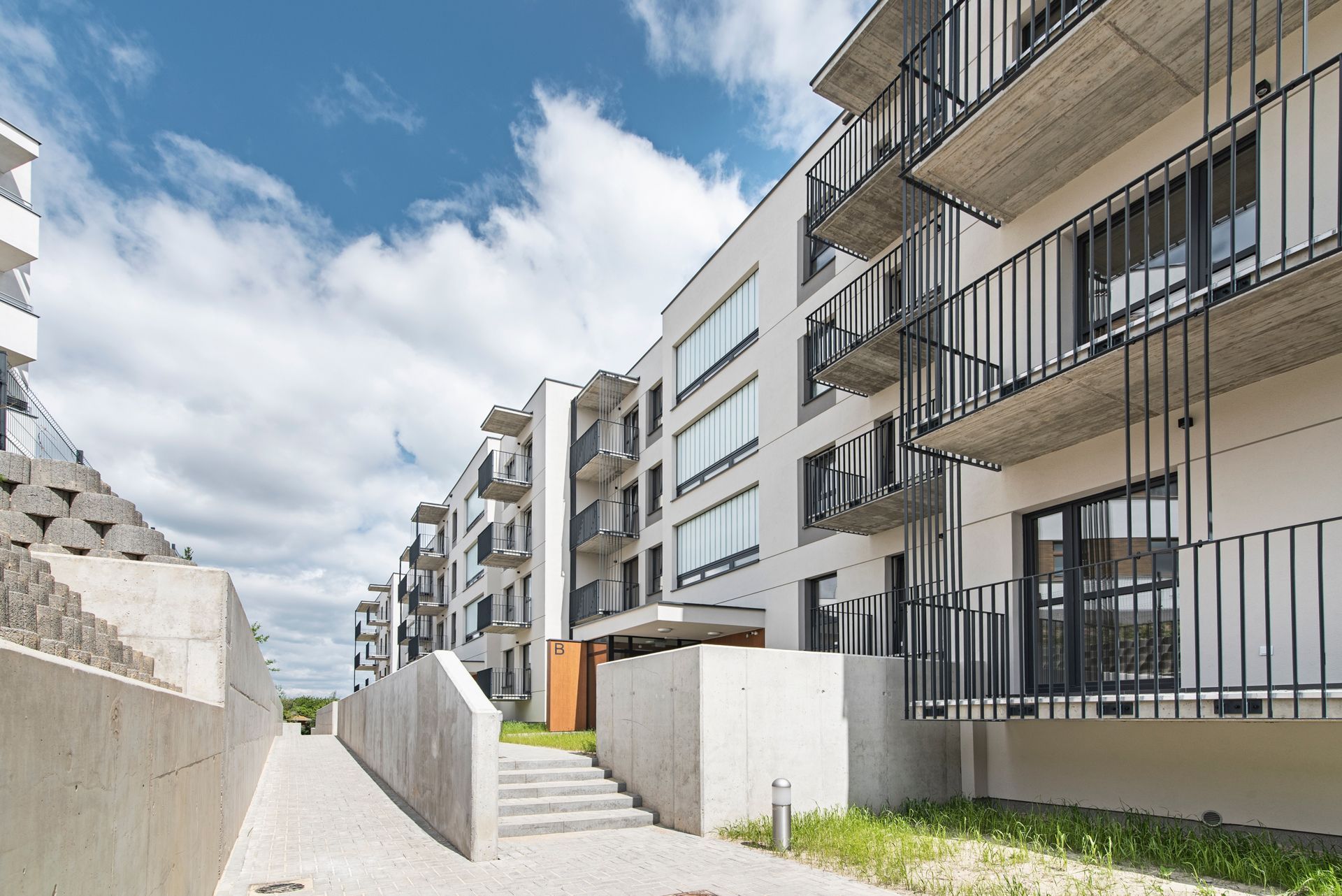Modern white apartment building with concrete balconies and a stone walkway under a bright, partly cloudy sky.