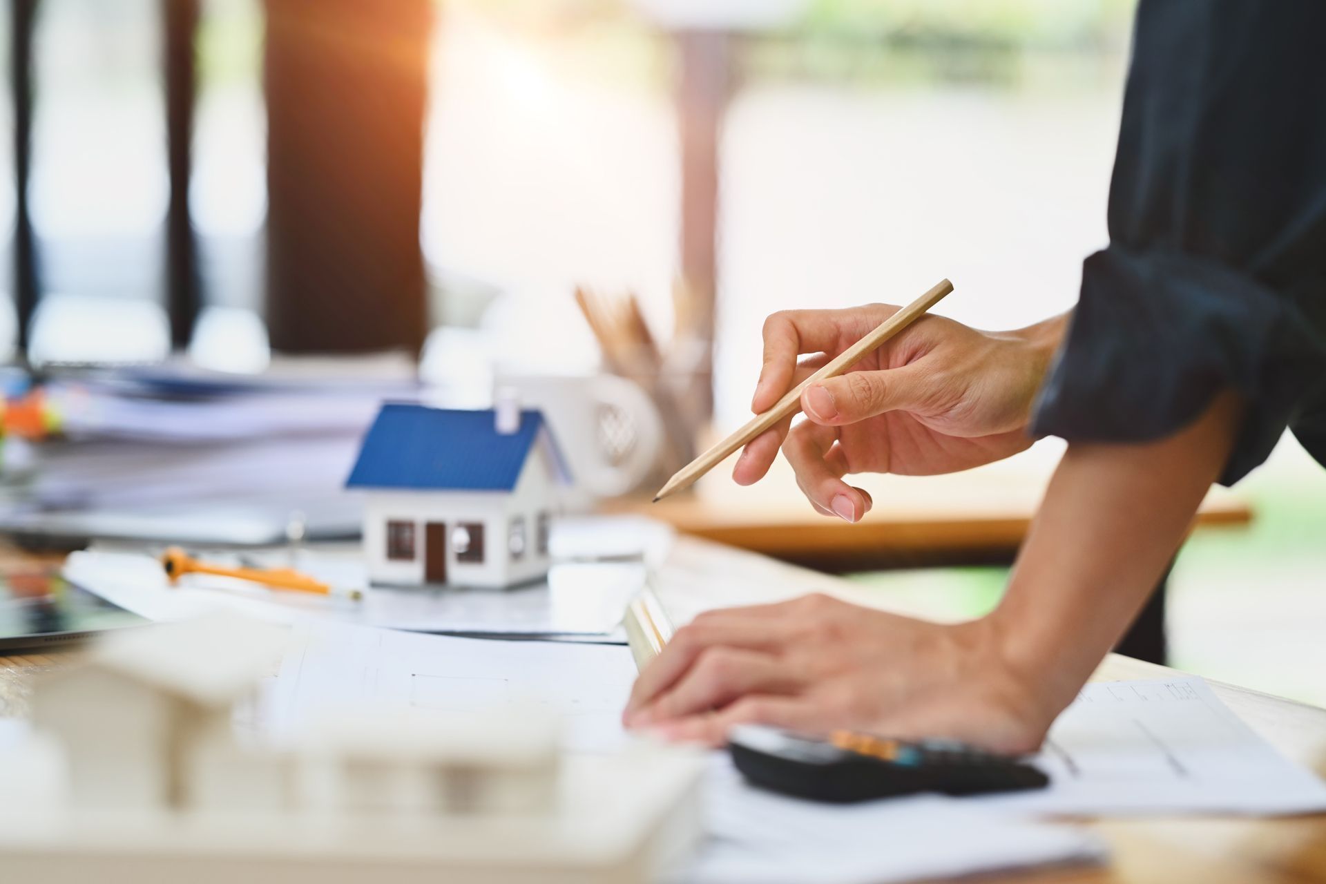 A person with a pencil works on building blueprints beside a small model house on a desk.