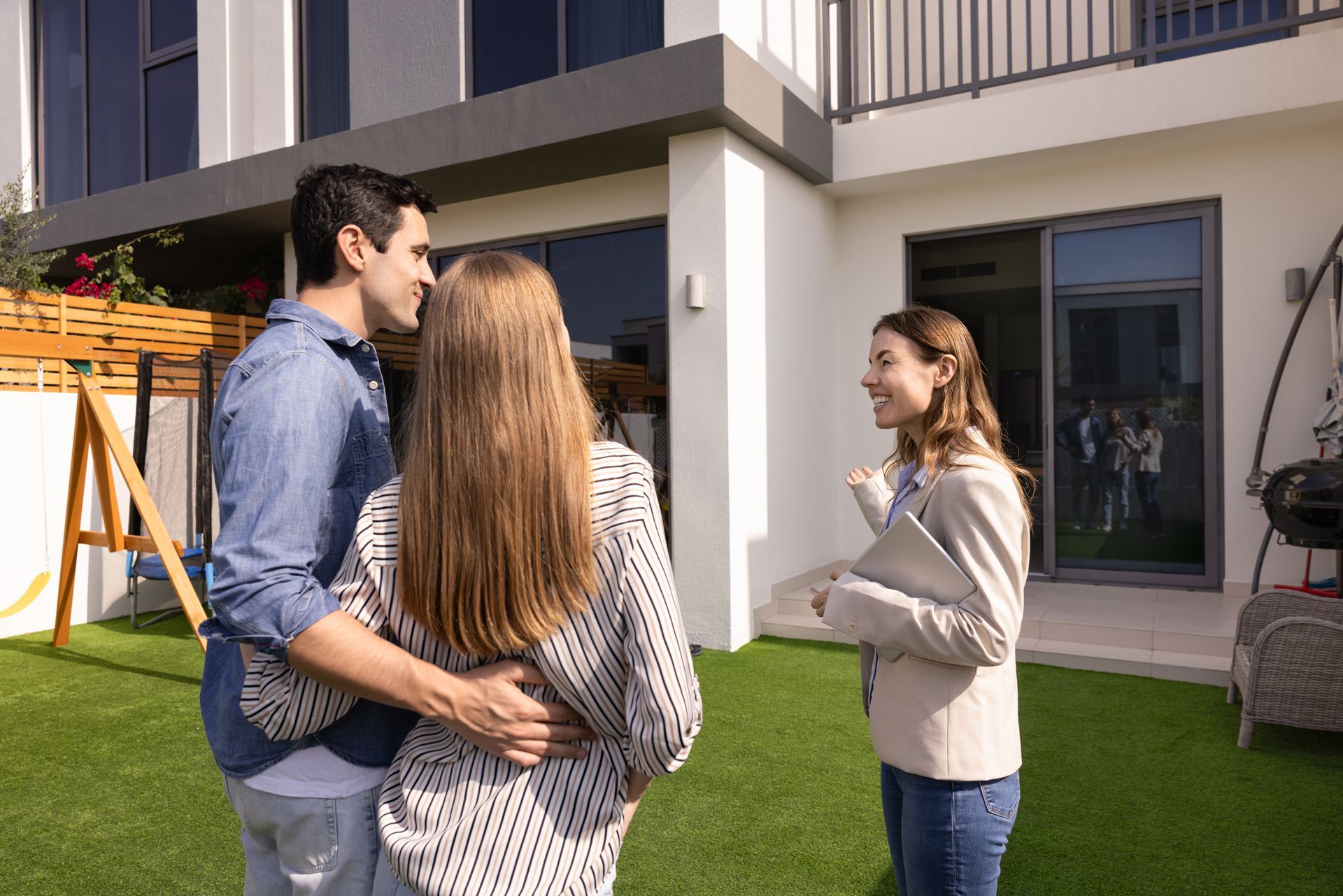 A real estate agent stands on a grassy patio, talking to a couple who are looking at a modern house.