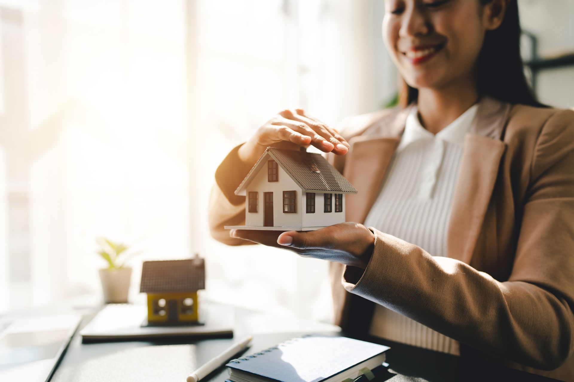 Woman holding a miniature house model beside another house on a table, in a bright office setting