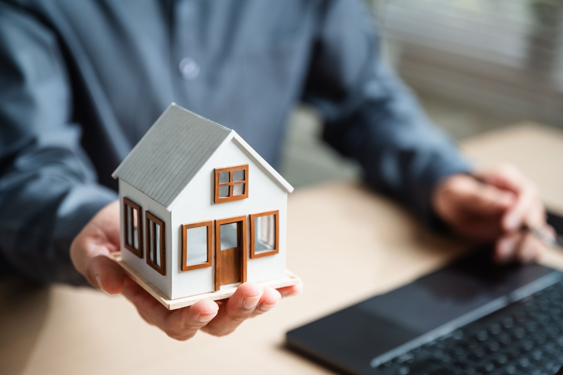 A person in a gray shirt holds a small model house in their hand over a desk with a laptop.