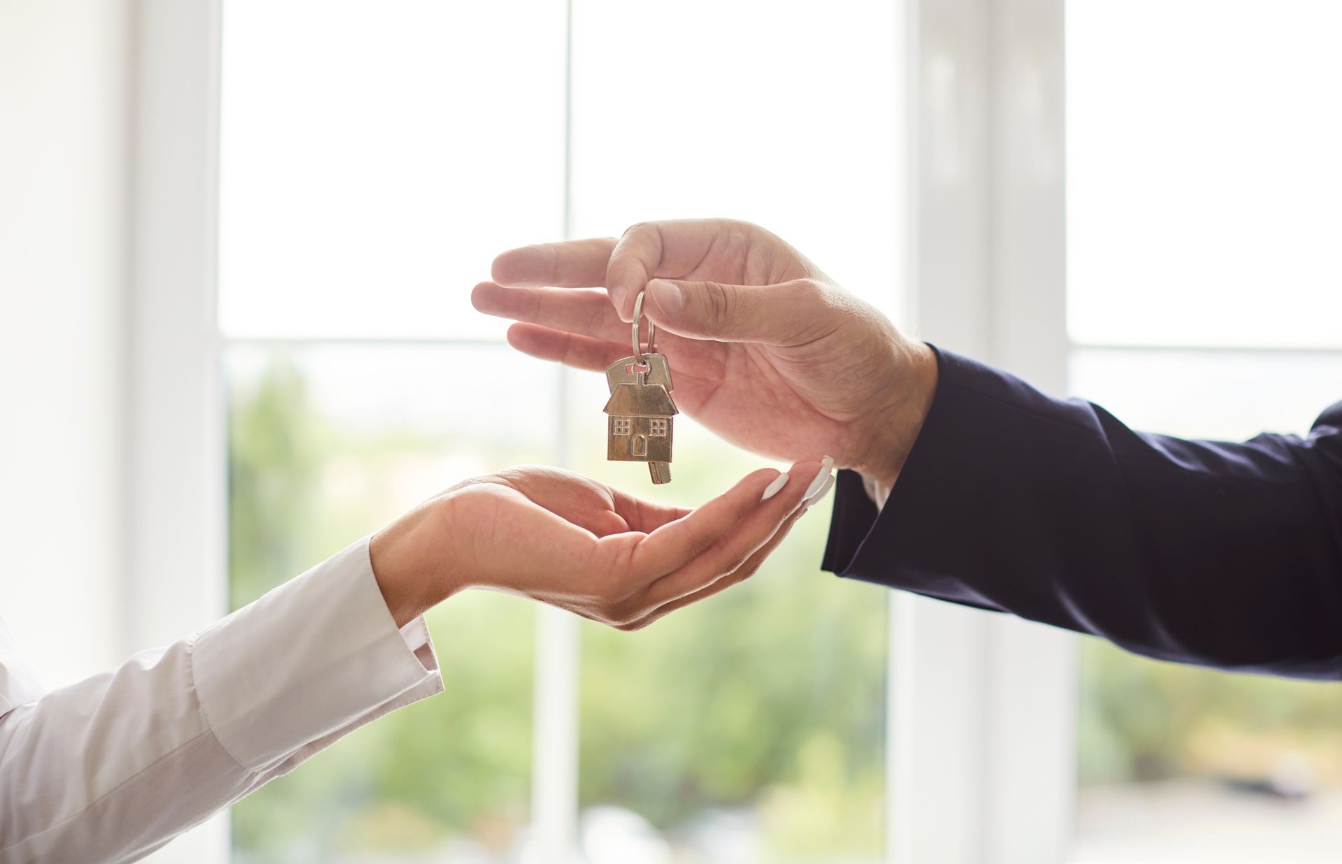 A person in a business suit handing a house-shaped key to another person's open palm in front of a bright window.