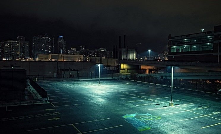 An empty parking lot is lit up at night with a city in the background.