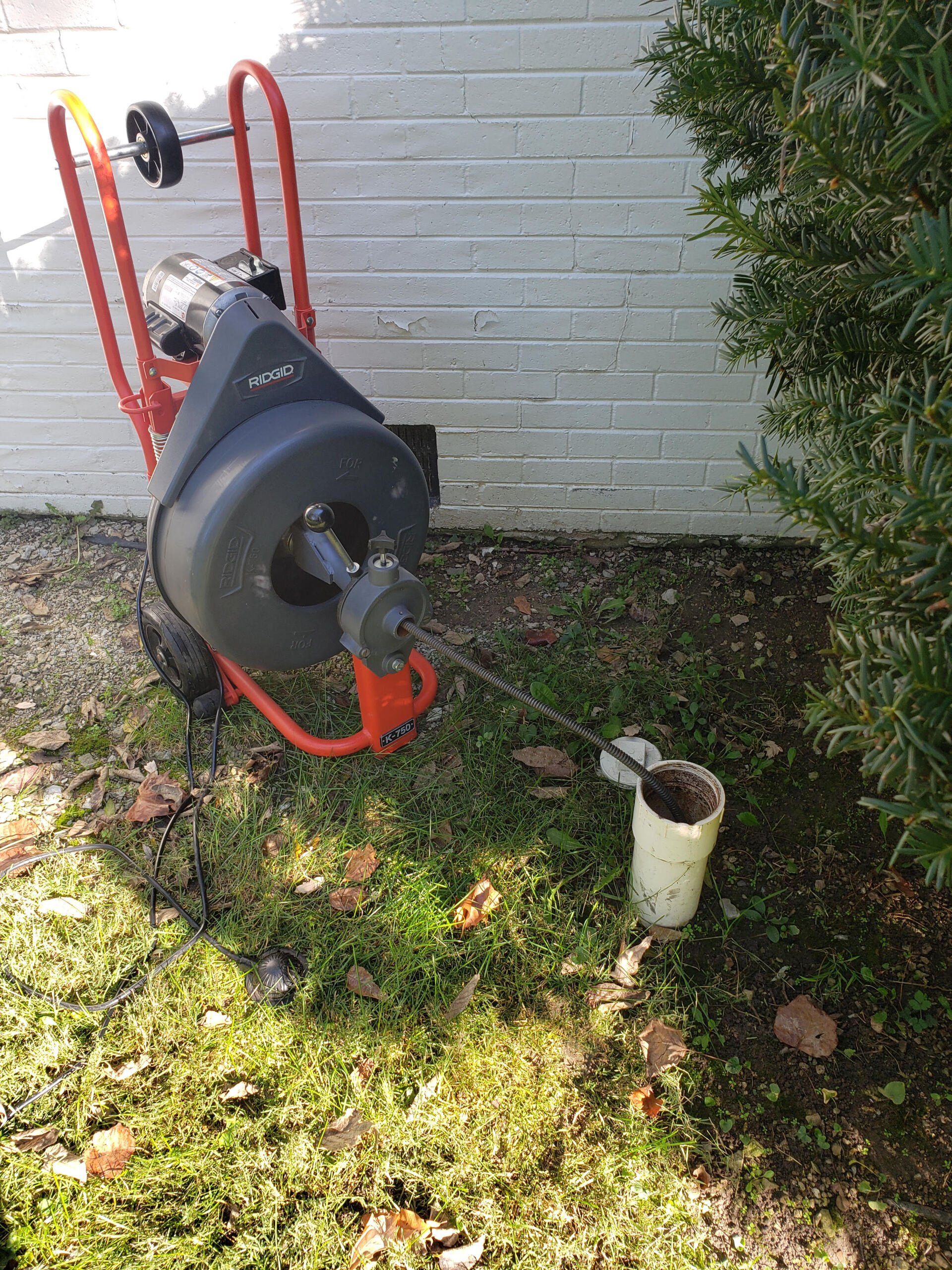 A drain cleaner is sitting in the grass next to a pipe.