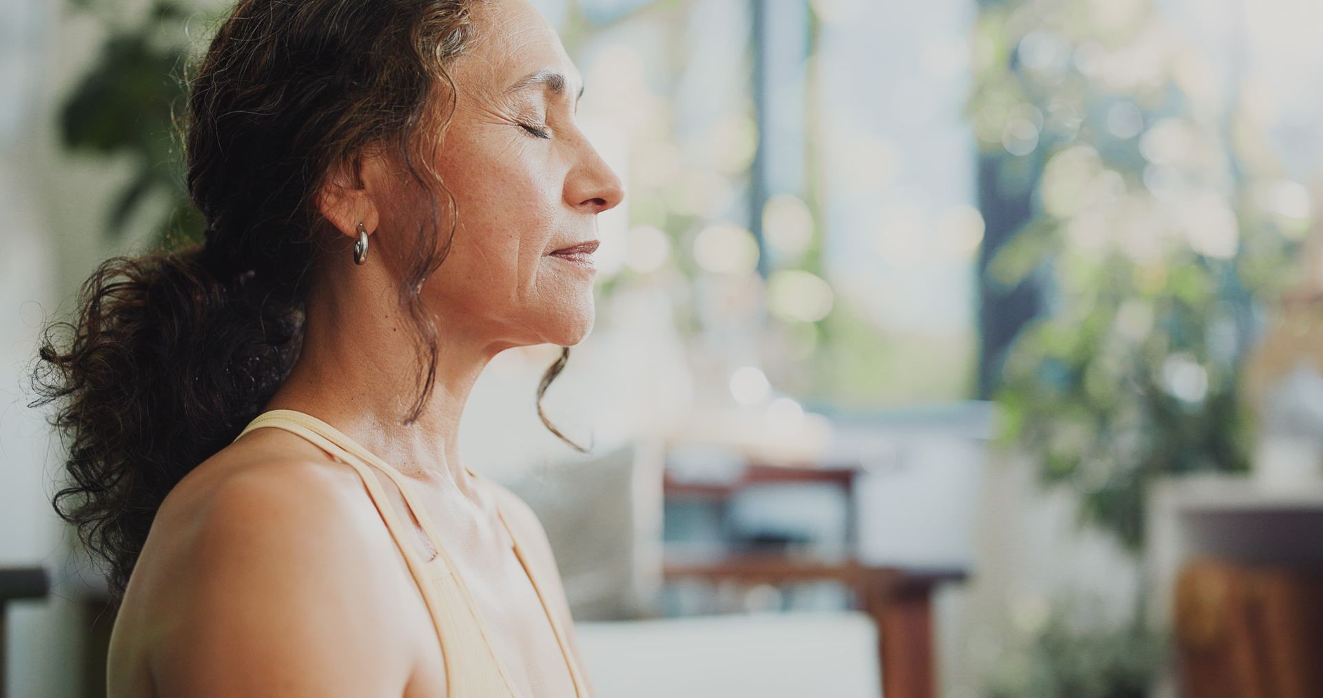 Woman with eyes closed, meditating indoors near plants; soft, natural light.