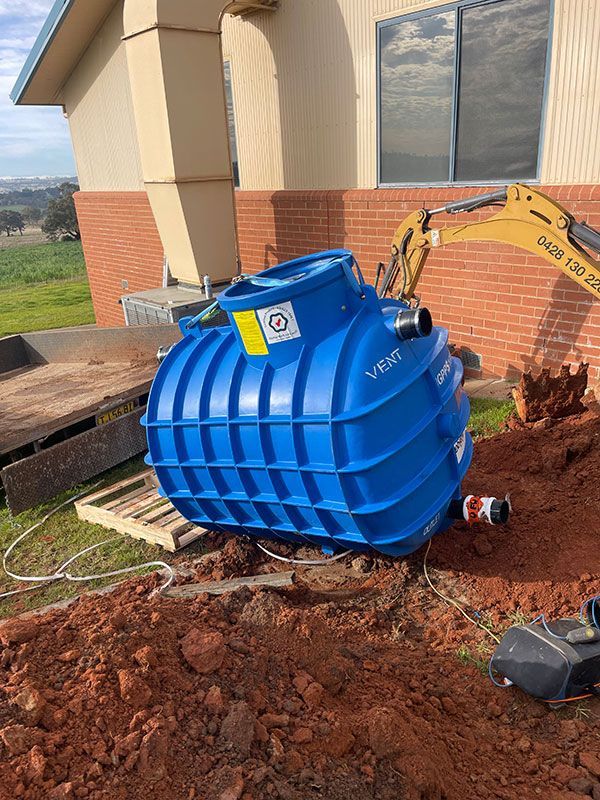 A Large Blue Tank Is Sitting In The Dirt In Front Of A Building — Pertzels Plumbing In Wagga Wagga, NSW