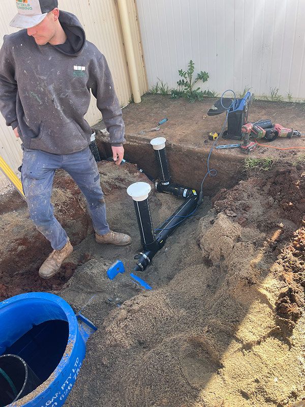 A Man Is Standing In The Dirt Next To A Blue Barrel — Pertzels Plumbing In Wagga Wagga, NSW