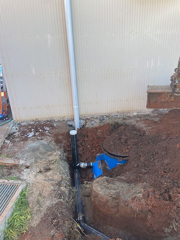 A Pipe Is Being Installed In A Hole In The Ground Next To A Building — Pertzels Plumbing In Wagga Wagga, NSW