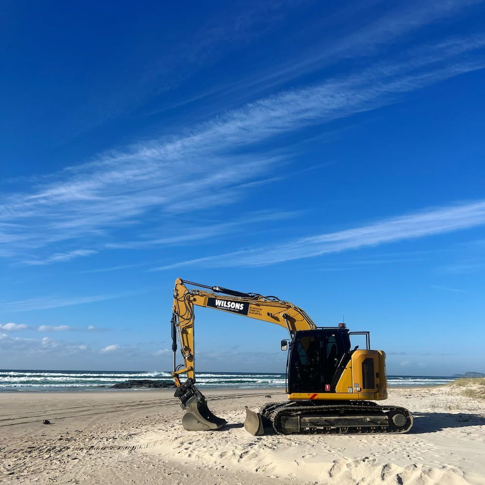 A Yellow Excavator Is Parked On A Sandy Beach — Wilsons Excavations & Plant Hire in Myocum, NSW