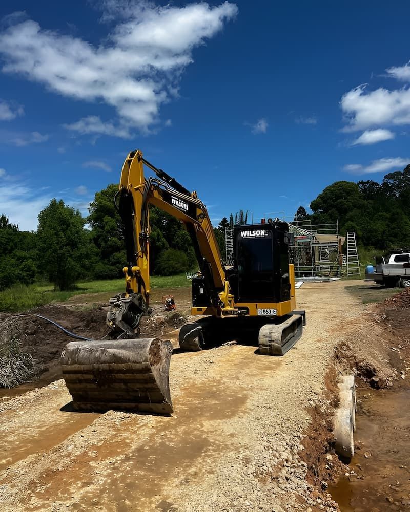 A Yellow And Black Excavator Is Driving Down A Dirt Road — Wilsons Excavations & Plant Hire in Mullumbimby, NSW