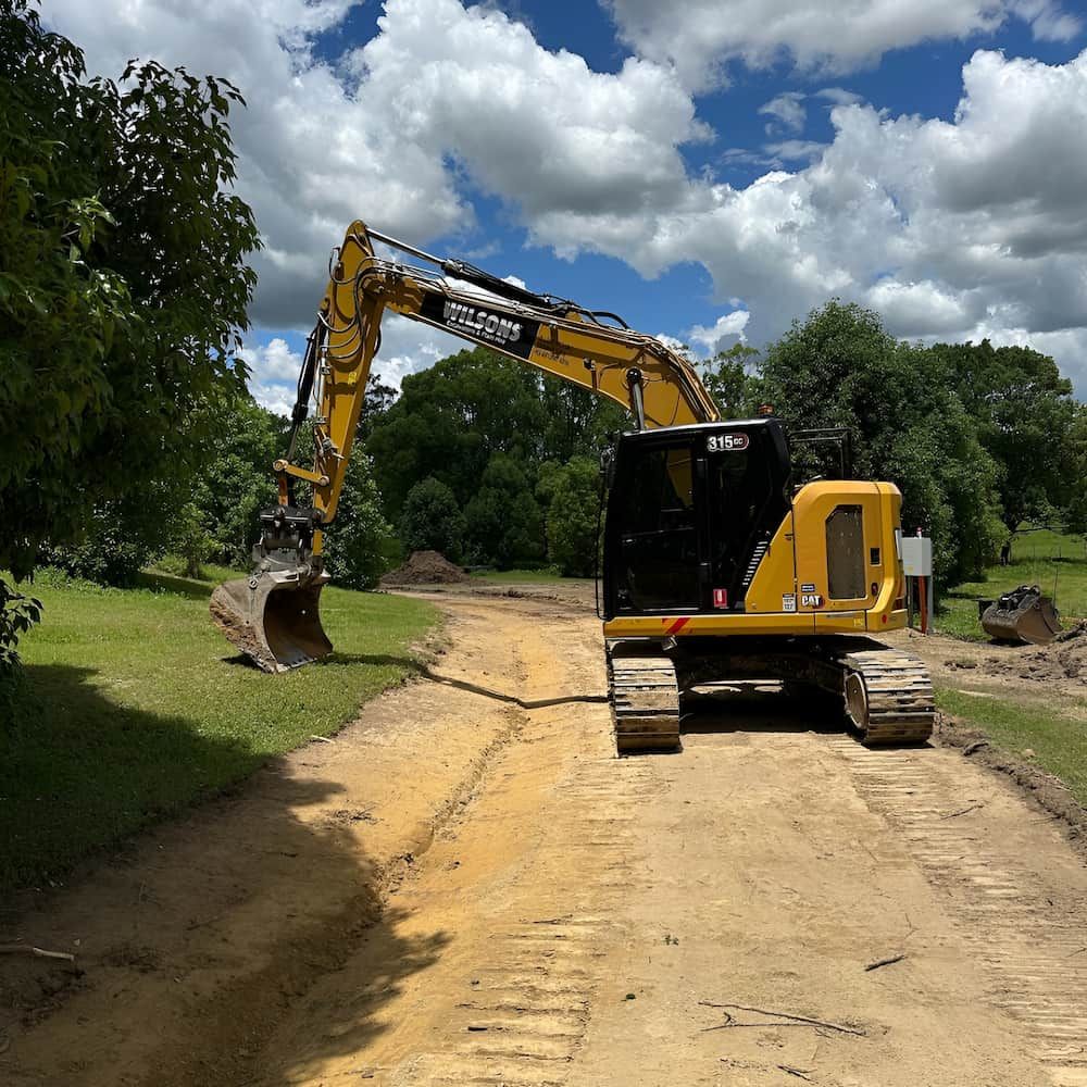 A Yellow Excavator Is Driving Down A Dirt Road — Wilsons Excavations & Plant Hire in Myocum, NSW
