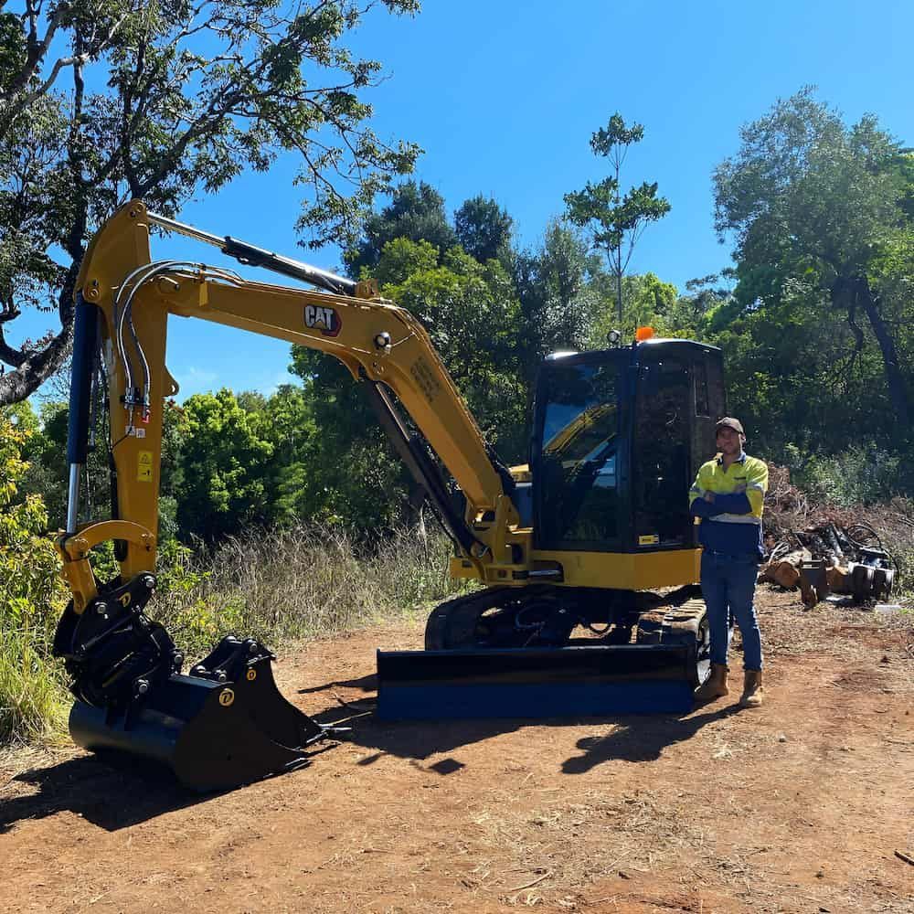 A Man Stands In Front Of A Yellow Cat Excavator — Wilsons Excavations & Plant Hire in Tweed Heads, NSW