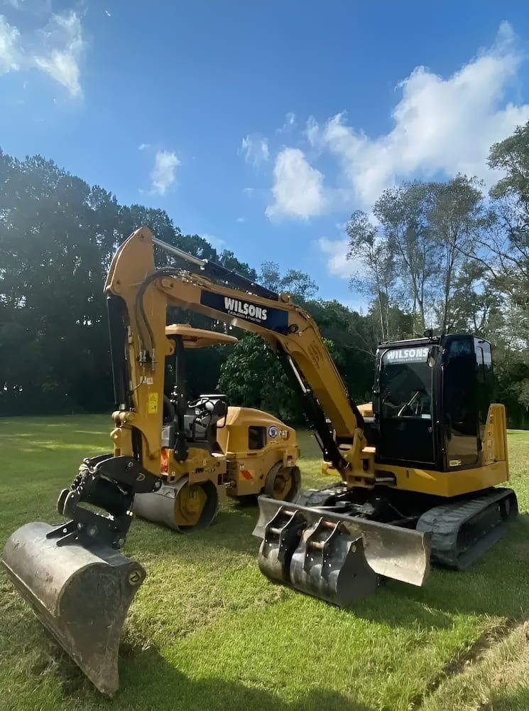 A Couple Of Excavators Are Parked In A Grassy Field — Wilsons Excavations & Plant Hire in Myocum, NSW