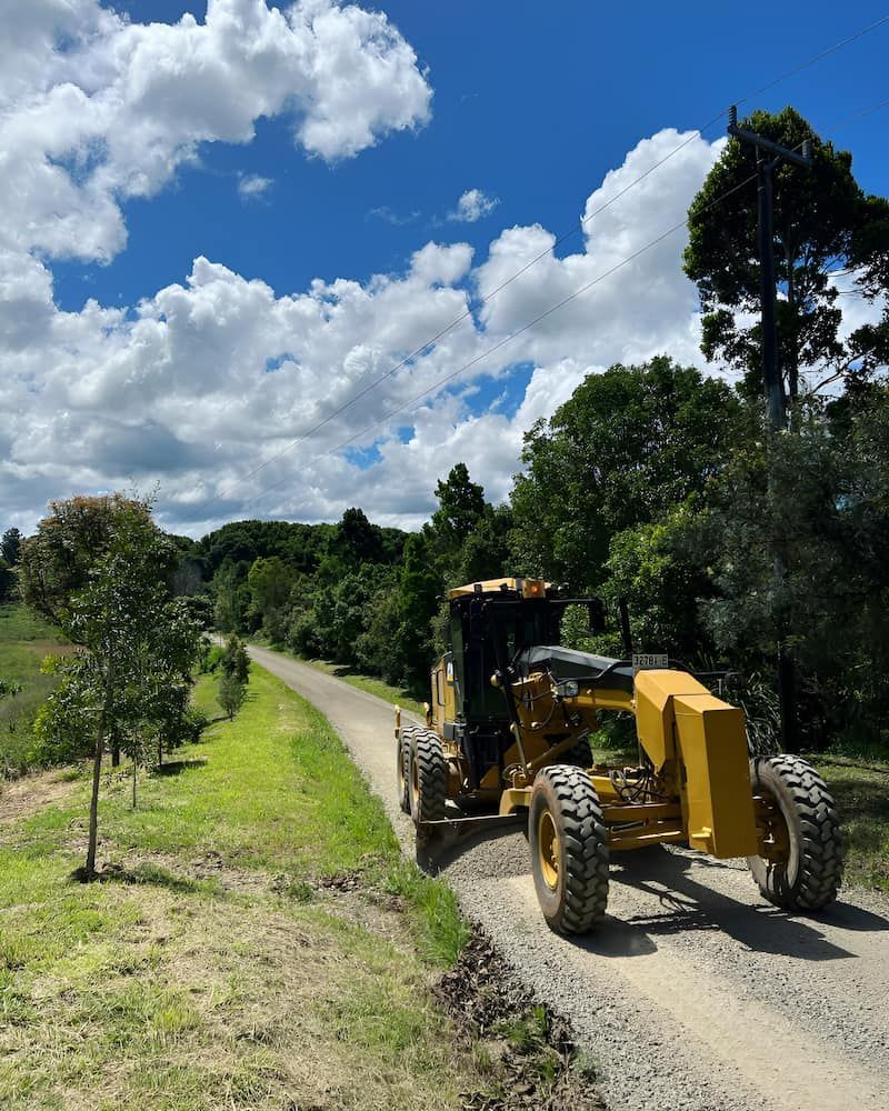 A Yellow Tractor Is Driving Down A Dirt Road — Wilsons Excavations & Plant Hire in Myocum, NSW