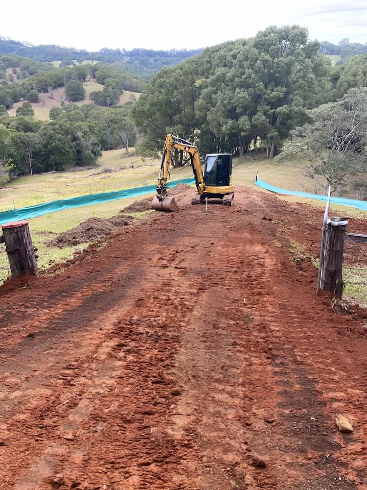 A Yellow Excavator Is Driving Down A Dirt Road — Wilsons Excavations & Plant Hire in Myocum, NSW
