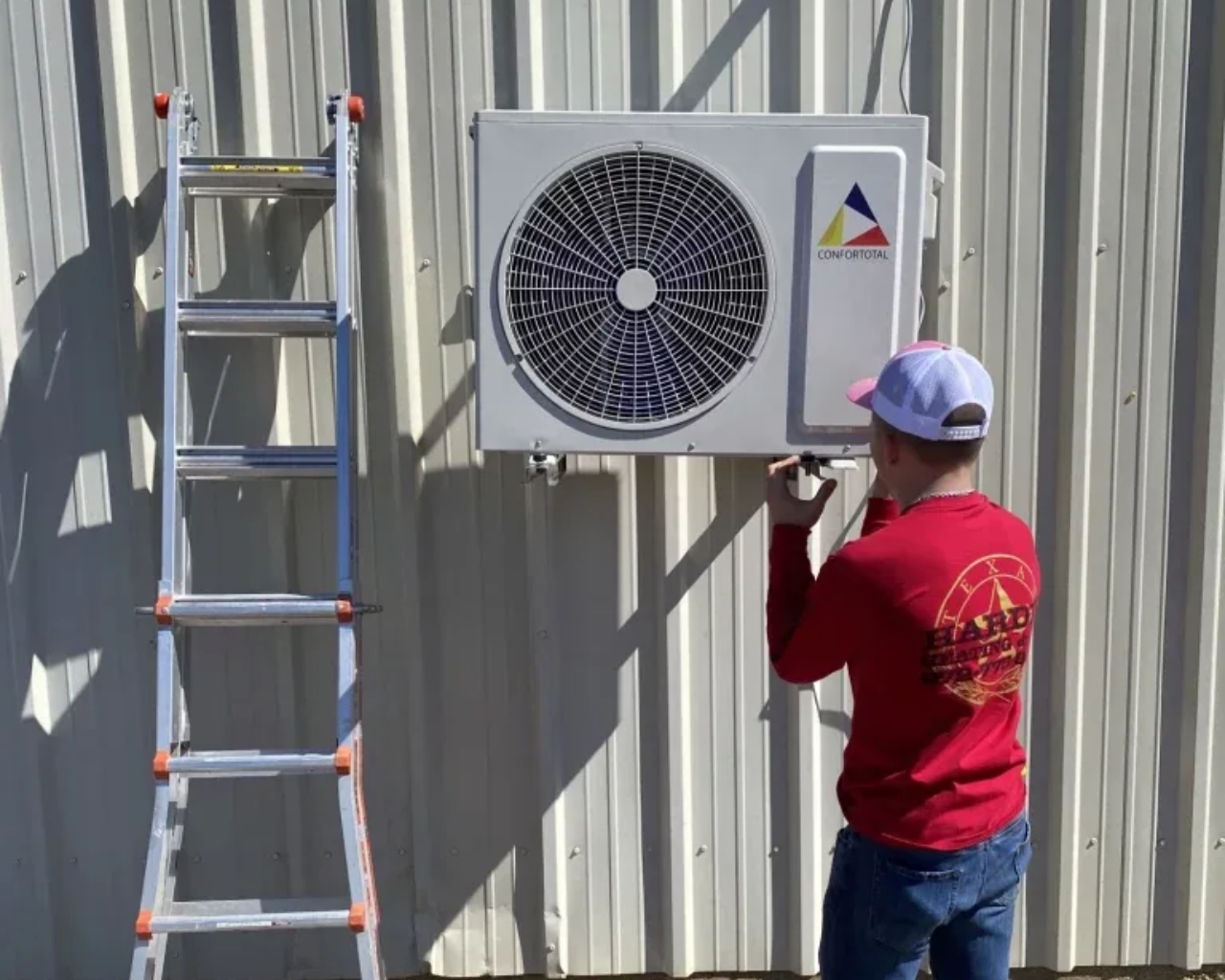 Person installing an air conditioner unit on a metal building. Ladder on the left.