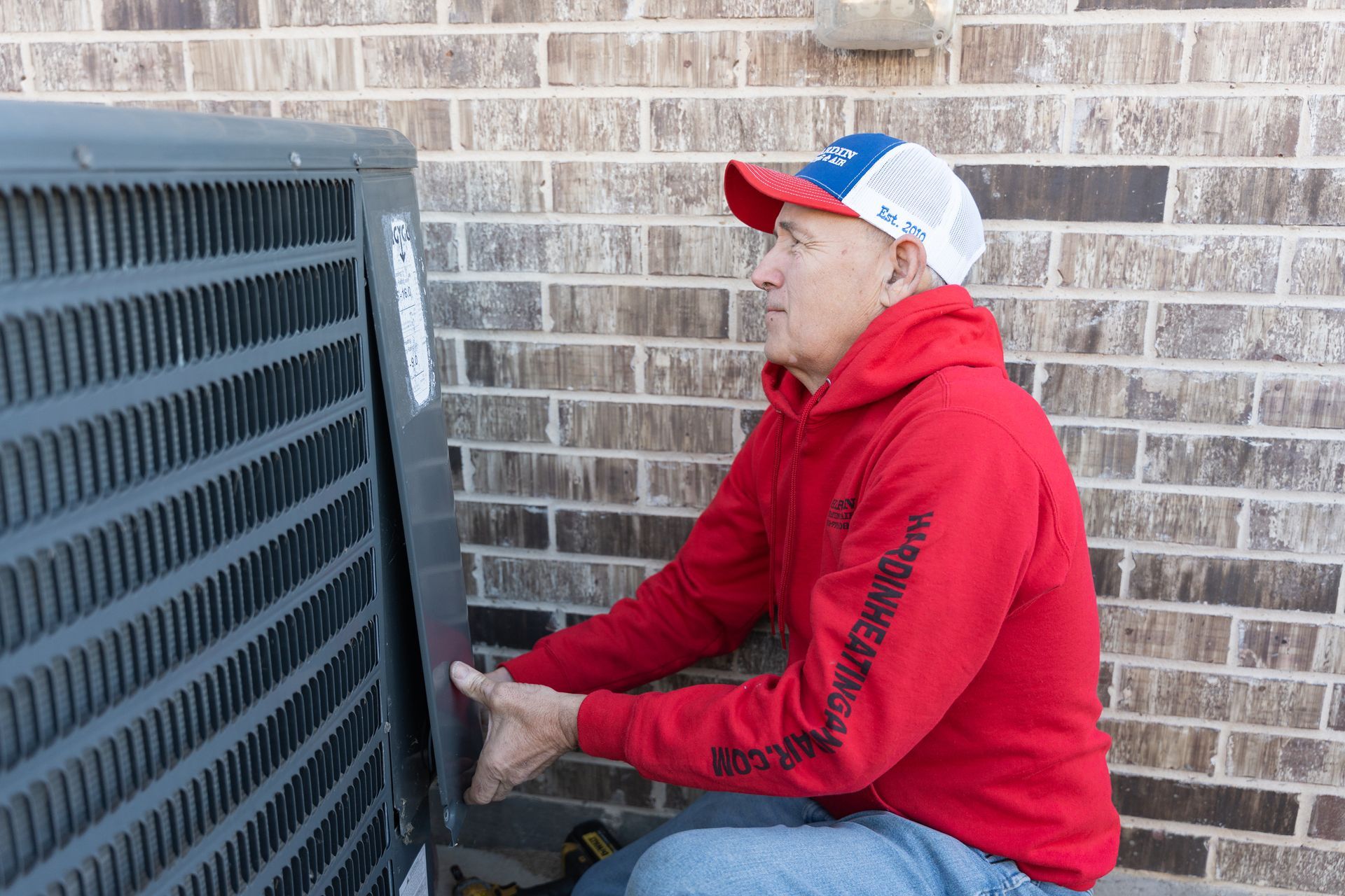Hardin Heating & Air Technician, cap removing panel from outdoor air conditioning unit next to brick wall.