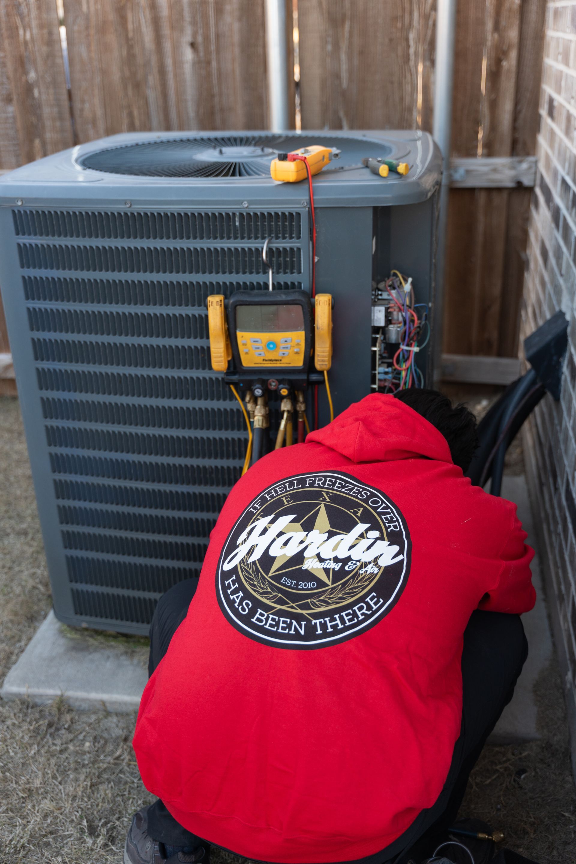 HVAC technician in red hoodie inspecting an air conditioning unit outside, with tools visible.