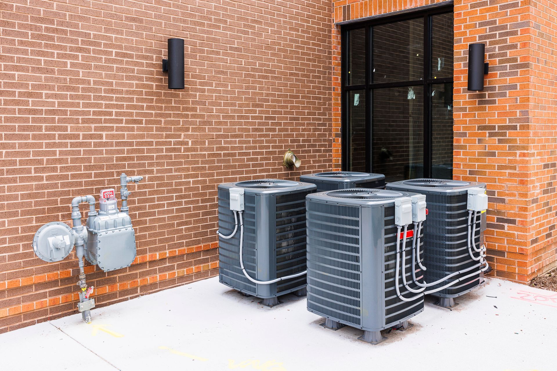 Air conditioning units against a brick wall next to a gas meter, with two sconces and a window.