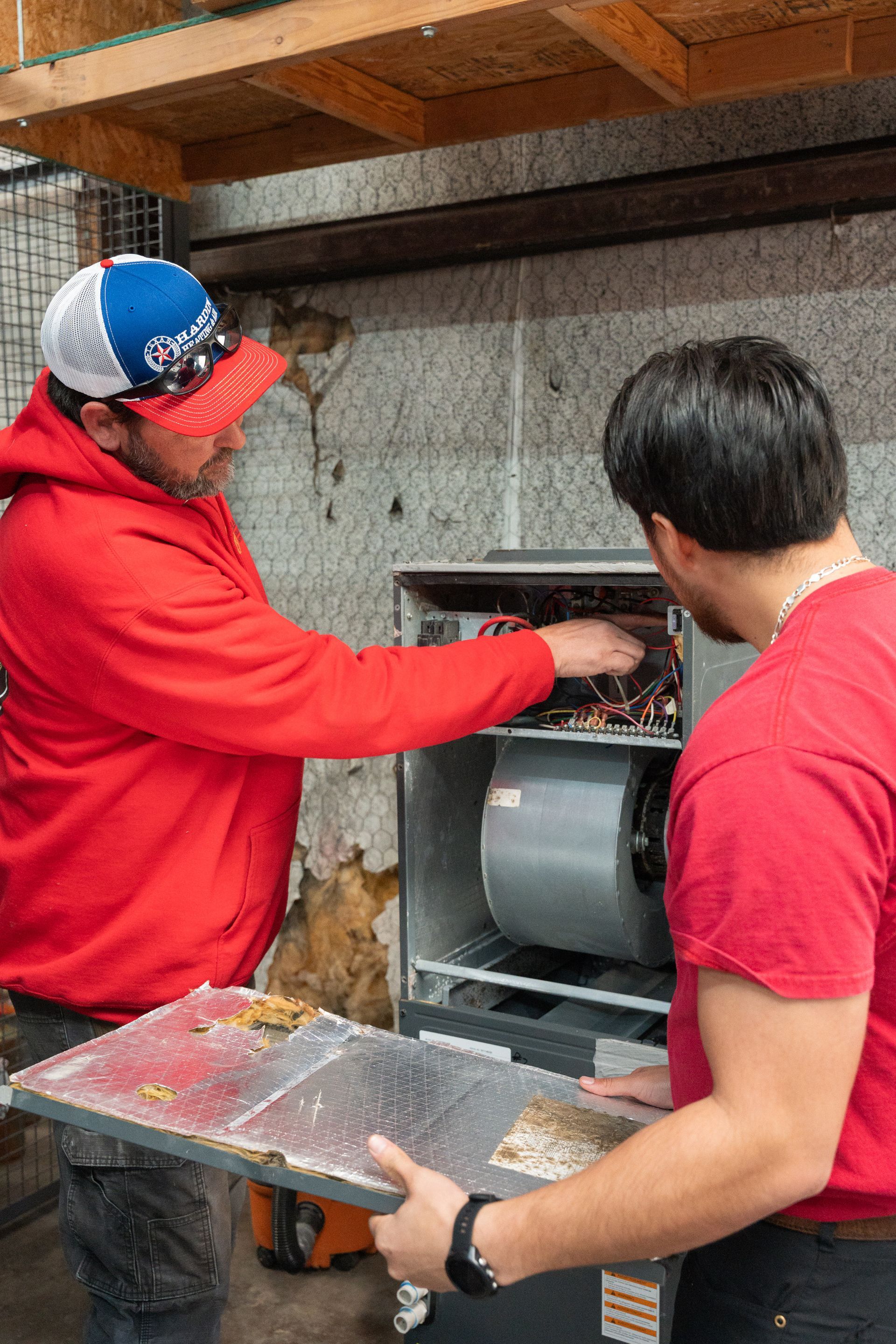 Hardin Heating & Air Technicians in red shirts inspect a metal appliance against a concrete wall.