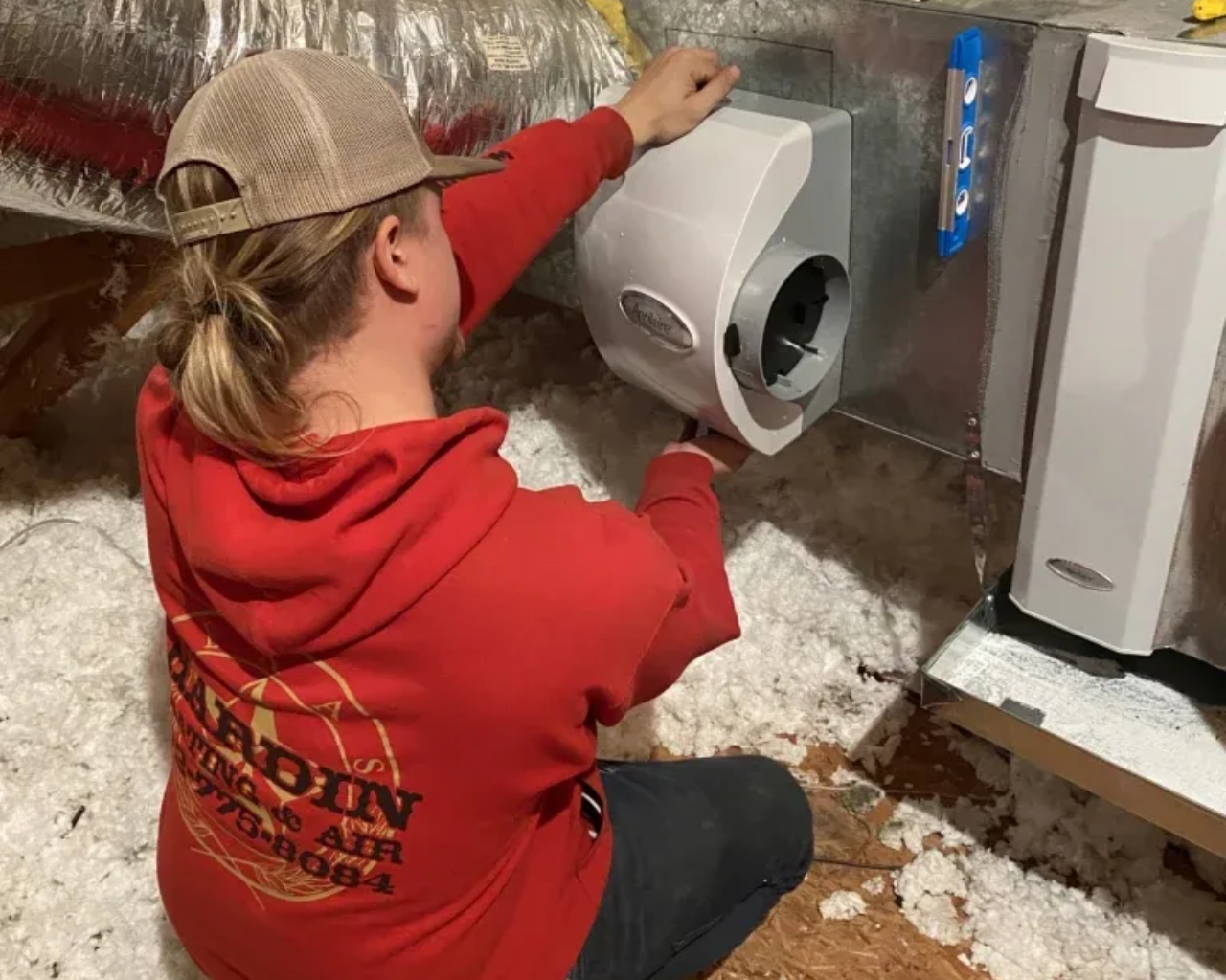 Hardin Heating & Air Technician installing a humidifier in ductwork, inside an attic. Wearing red hoodie and tan cap.