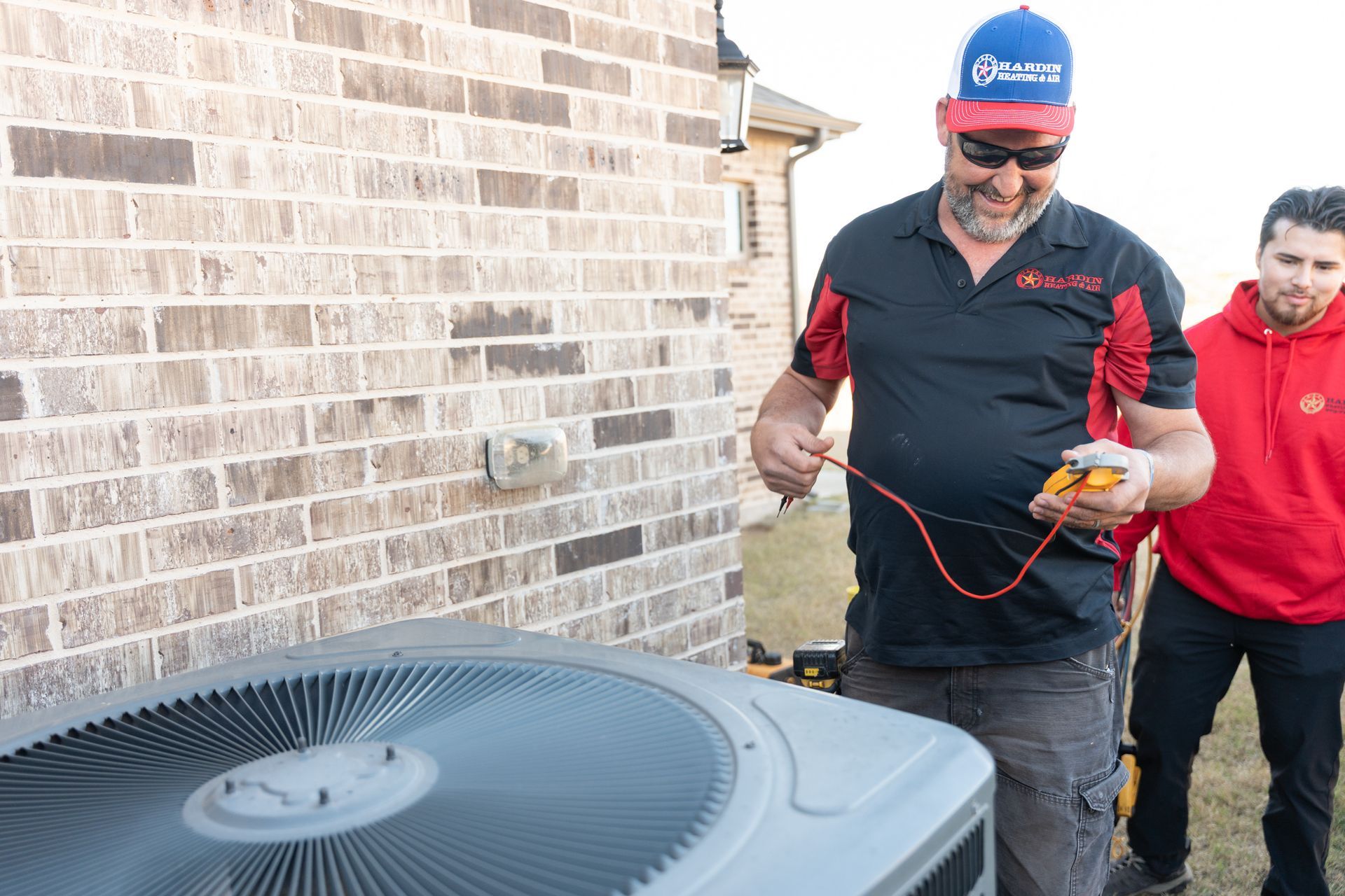 Two men inspecting an AC unit near a brick building. One holds a multimeter; the other watches.